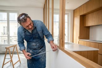 Artisan inspectant une verrière intérieure en bois mal assemblée entre salon et cuisine dans un appartement parisien